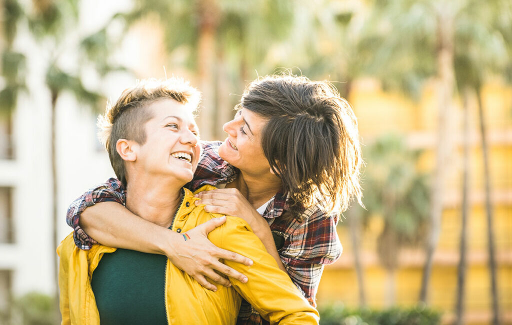 Woman and nonbinary person in a relationship embracing outdoors. Woman embracing her partner. They are looking back at her smiling.