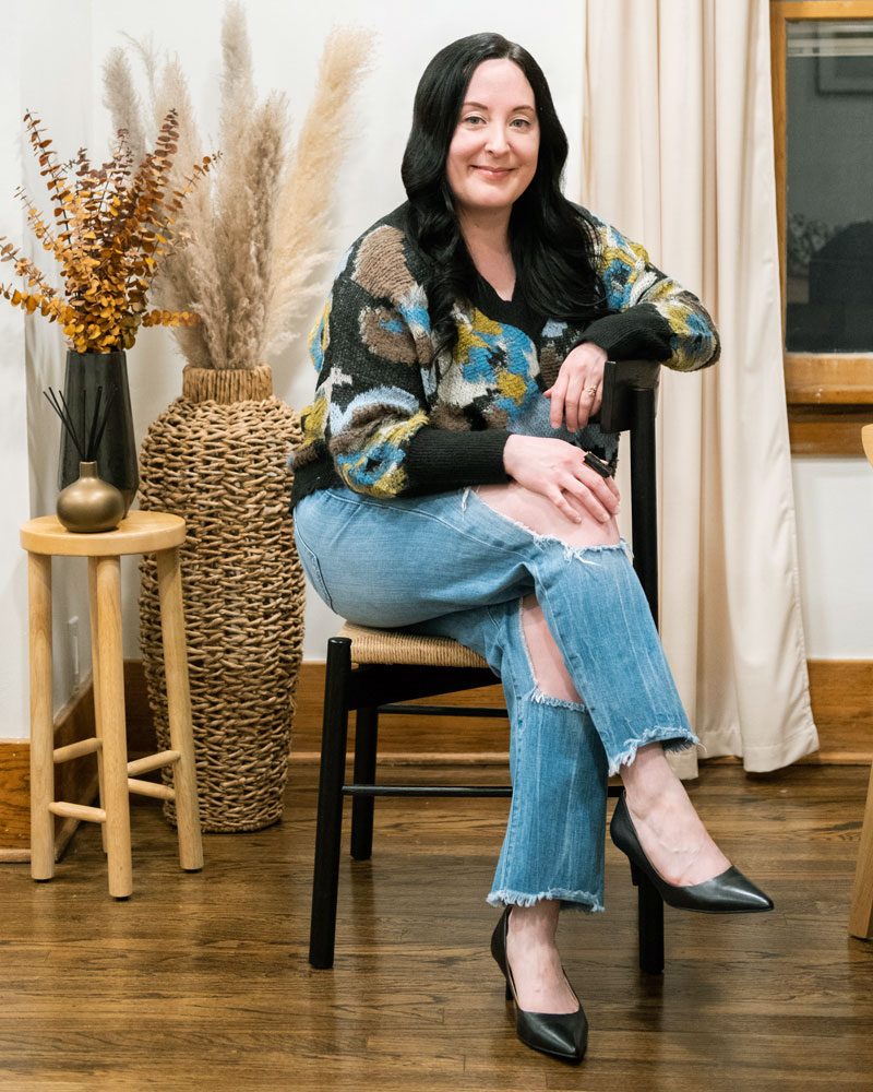 Alexis Buhrman sitting in a chair in a living room with her legs crossed, smiling at the camera