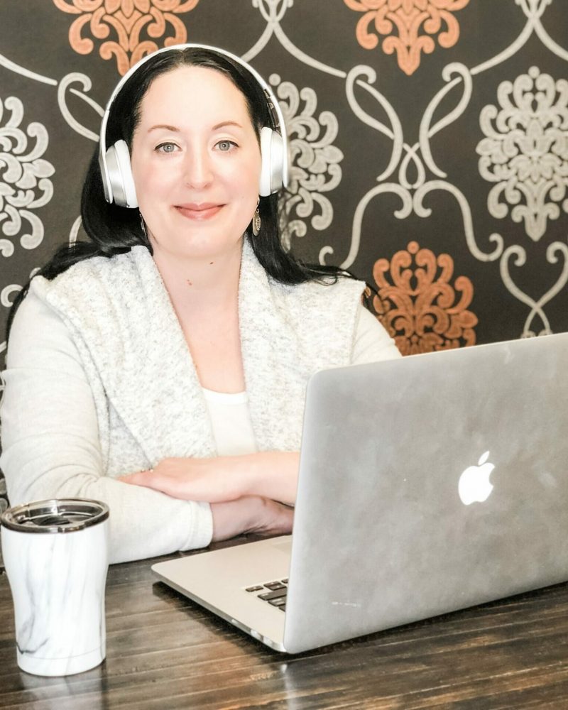 Alexis Buhrman sitting at a table with a laptop computer. Her arms are corssed and she's wearing headphones, smiling at the camera.